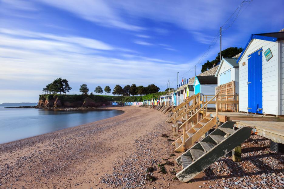 Beach huts lining Corbyn Beach in Torquay, with calm water and a blue sky.
