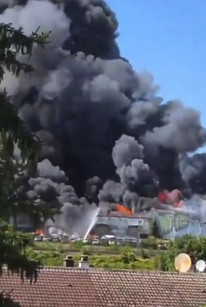 A large fire with thick black smoke billowing into the sky above a building and surrounding vehicles, with firefighters spraying water.