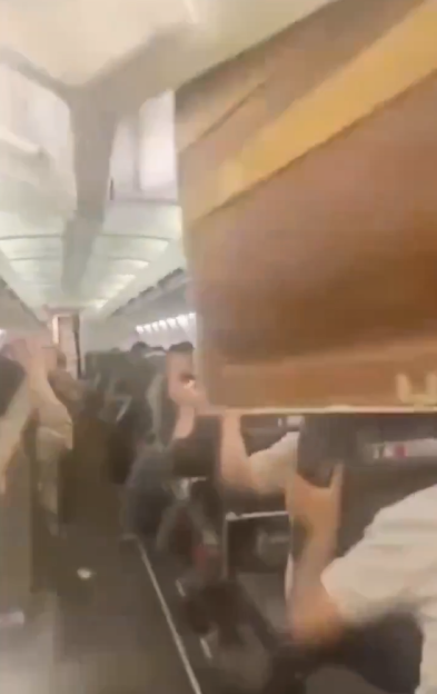 Passengers ducking as an overhead compartment in an airplane cabin falls open, spilling contents.