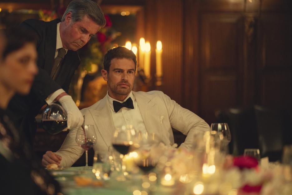 A waiter pours red wine for a man in a cream tuxedo at a candlelit table.