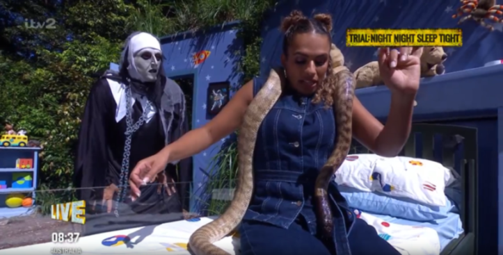 Woman with a snake draped around her neck and shoulders, while a person in a ghost costume stands beside her.