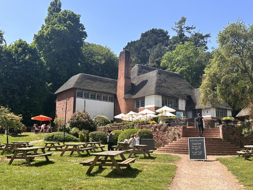 A large thatched-roof building with outdoor seating, picnic tables, and lush green landscaping.
