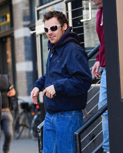 Harry Styles wearing a navy jacket and blue jeans, walking outside while holding a phone.