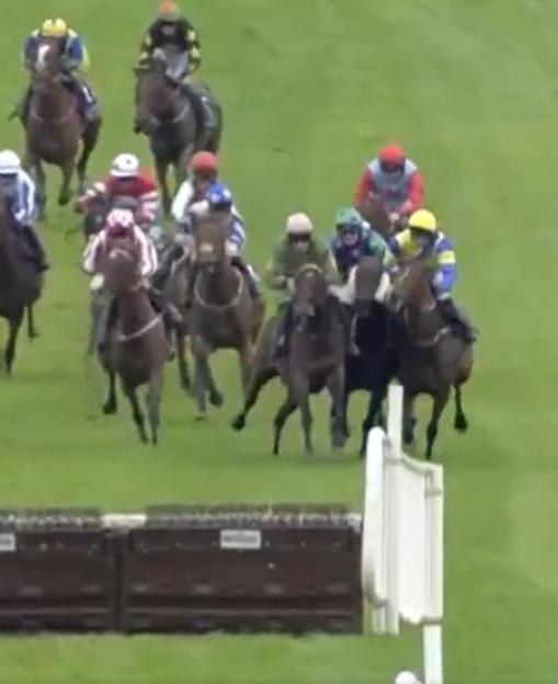 Horses and jockeys racing past a jump hurdle on a grass track.