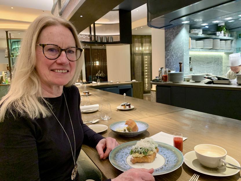 A woman in glasses smiles while seated at a restaurant bar with a chef in the background.