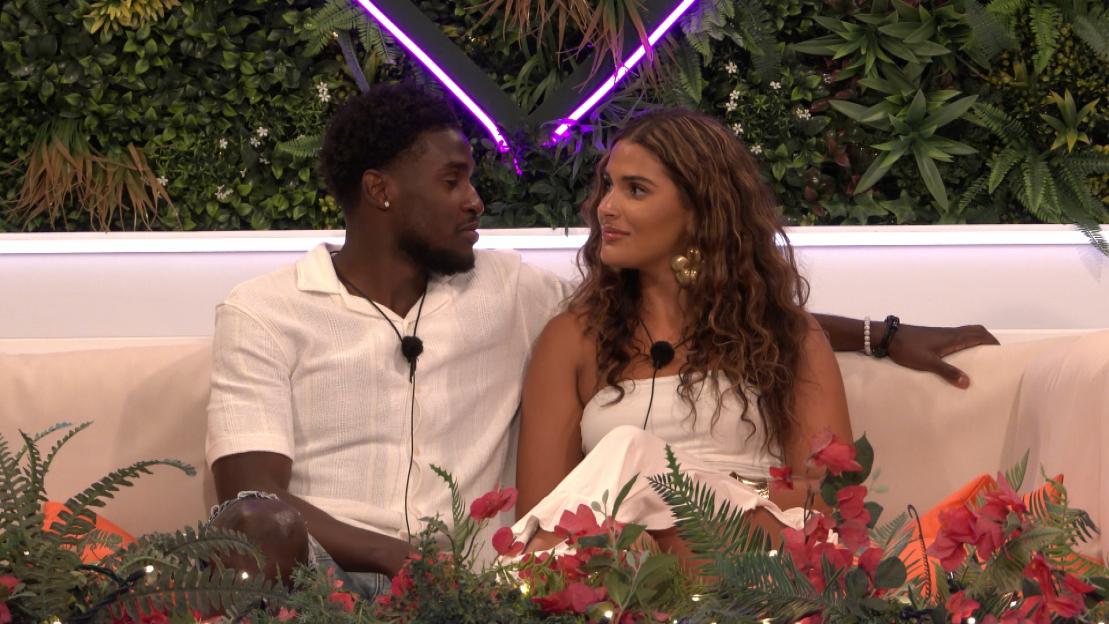 Mica and Simba from Love Island sitting on a sofa with lush green foliage and neon lights in the background.