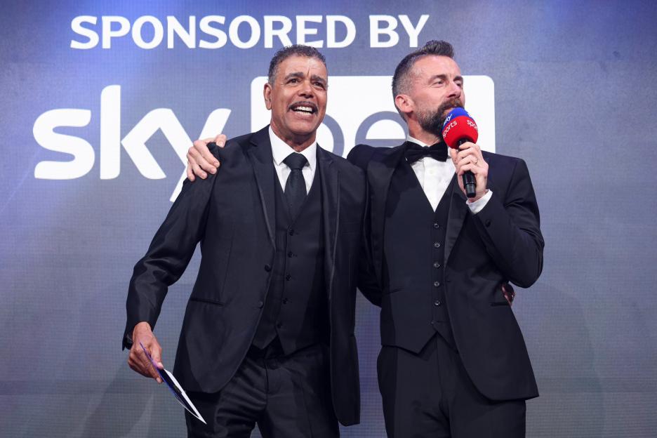 Two men in suits, one holding a microphone, standing on stage in front of a "Sponsored by Sky" banner.
