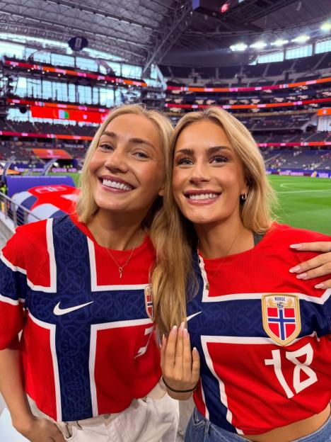 Two women wearing matching red and blue shirts with the Norwegian flag and emblem, smiling inside a stadium.