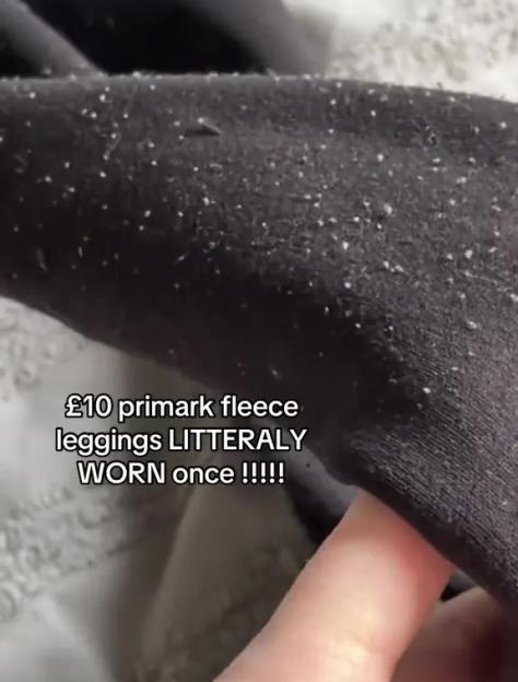 Close-up of a hand stretching out a piece of black fleece fabric covered in white lint and pills, with text indicating it's a £10 Primark fleece worn once.