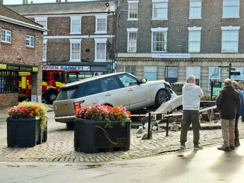A silver Range Rover has mounted a paved area and knocked down the Market Cross, with onlookers watching the scene and a fire engine parked in the background.