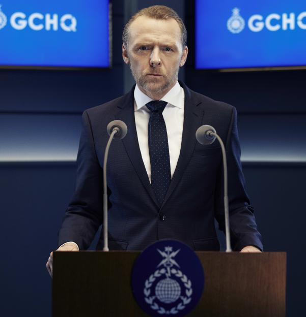 Simon Pegg, as Danny, stands sternly behind a podium with GCHQ logos visible.