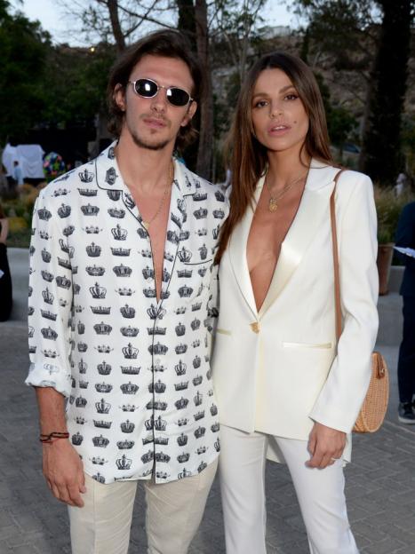 Man in a crown-patterned shirt and sunglasses posing with a woman in a white suit.