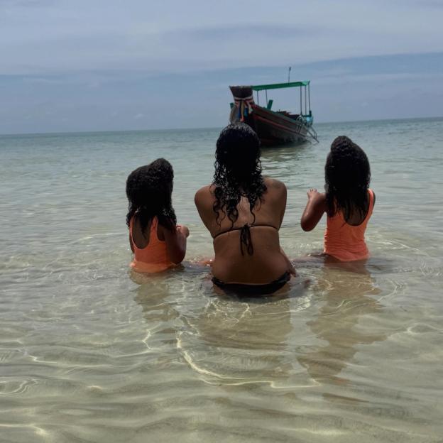 Three people, a woman and two children, sitting in clear shallow water looking out at a boat and the ocean.