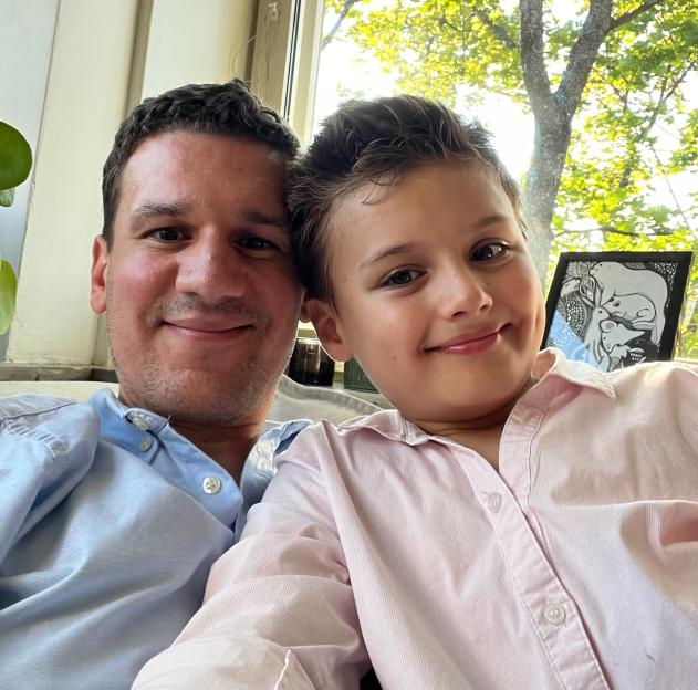 A man in a blue shirt and a boy in a pink shirt smiling at the camera, with a window and green trees in the background.