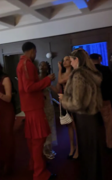 Several people in formal wear gather in a room with blue and red lighting, a man in a red suit talking to a woman in a fur coat.