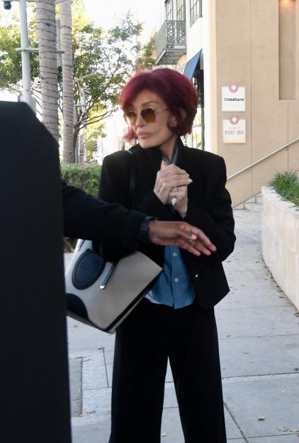 Sharon Osbourne wearing a black blazer, blue button-down shirt, wide-leg trousers, oversized sunglasses, and a Dior tote bag.