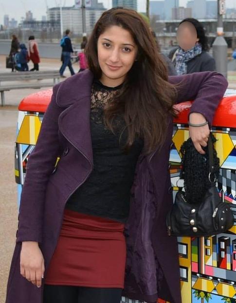 A young woman with long brown hair, wearing a purple coat and red skirt, leaning on a colorful structure outdoors.