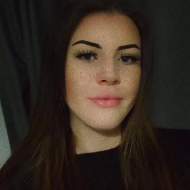 A young woman with long brown hair, prominent eyebrows, and freckles looks directly at the camera.