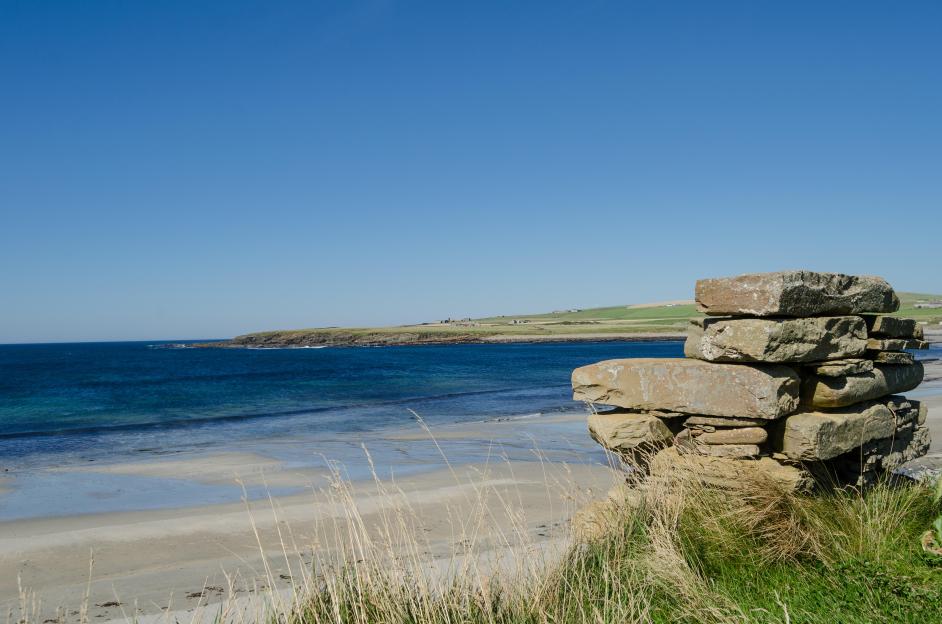 A sandy beach and blue ocean, with a green, hilly coast and scattered buildings in the distance, and a stacked stone wall in the foreground.