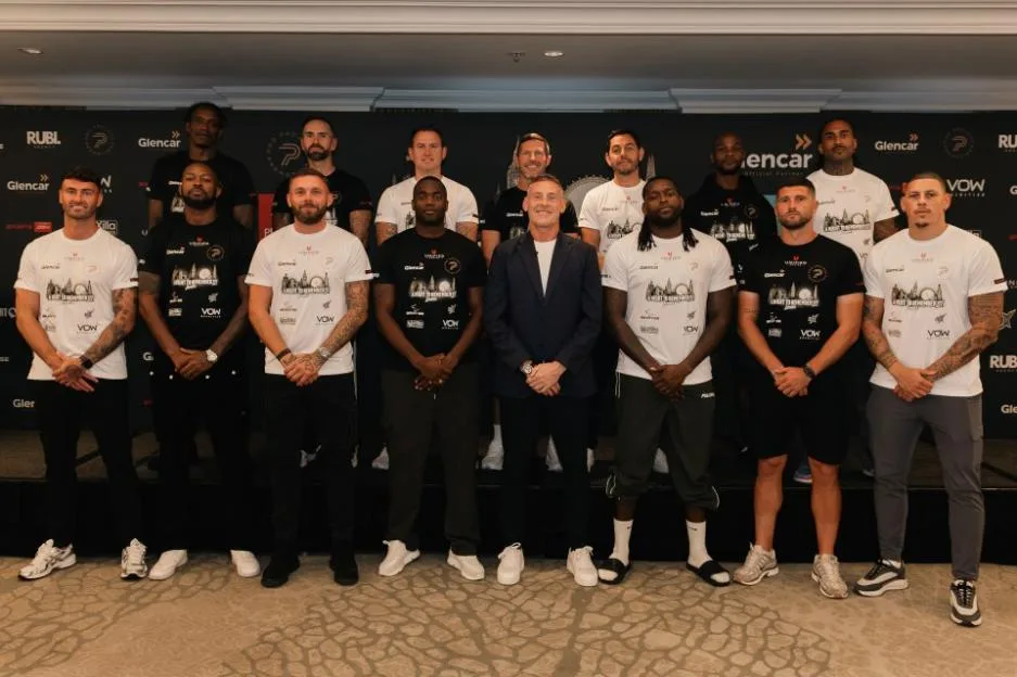 Group photo of sixteen men posing in two rows in front of a sponsor backdrop, with one man in the center of the front row wearing a suit.