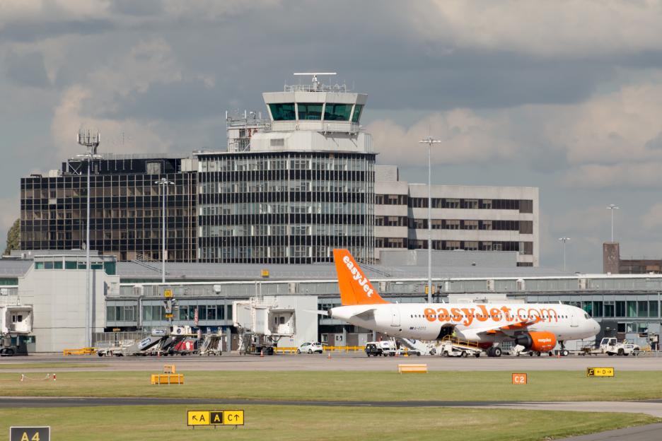 easyJet Airbus A319 passenger plane parked at Manchester Airport terminal with air traffic control tower in the background.