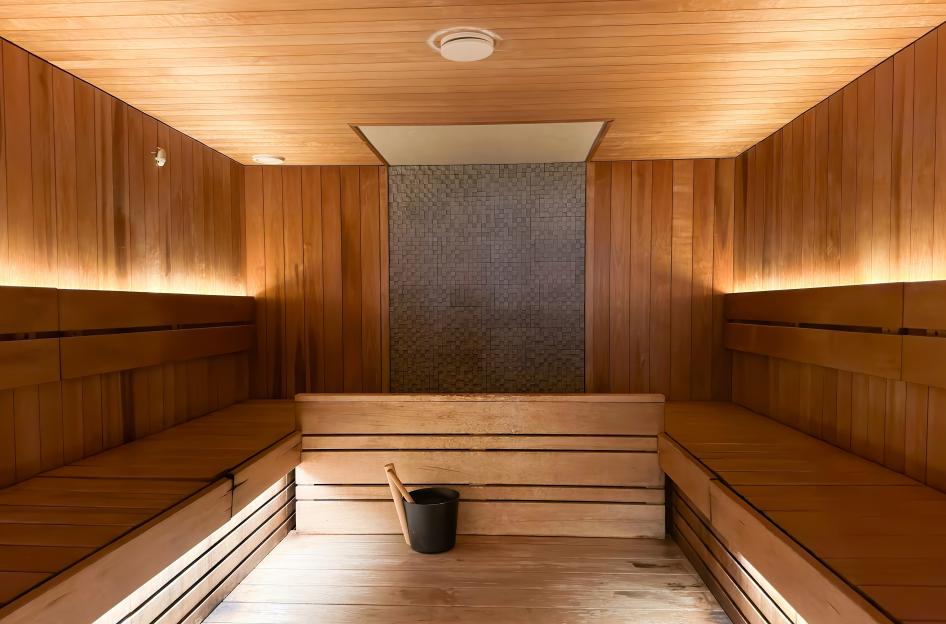 Modern Finnish sauna room with warm wooden walls and benches, ambient lighting, and a bucket and ladle on the floor.