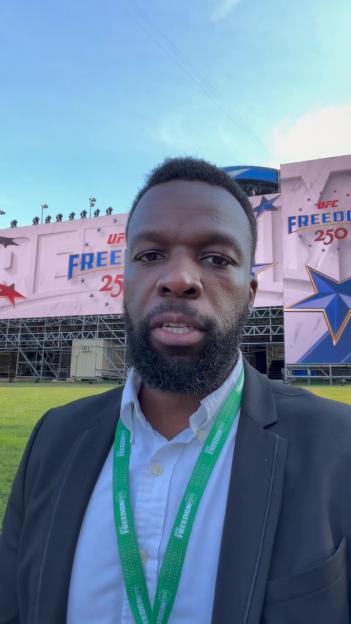 A man in a suit and white shirt with an "UFC Freedom 250" lanyard speaking in front of a stage decorated with "UFC Freedom 250" banners.