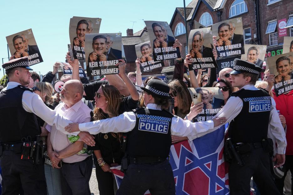 Police officers separate protesters holding anti-Keir Starmer signs, some displaying Starmer holding a mask of a grinning face over his real face, and a "STOP THE HATE" banner.