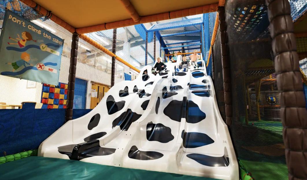 Four children sliding down a black and white cow-patterned slide in an indoor play area.