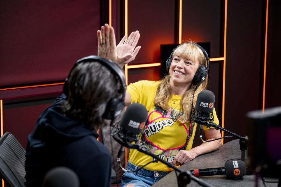 Sara Cox wearing a yellow "Pudsey" shirt and headphones, giving a high-five in a radio studio.