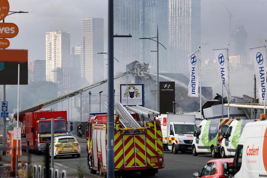 Firefighters spraying water on a burning commercial building, with fire trucks, police cars, and other service vehicles on the street.