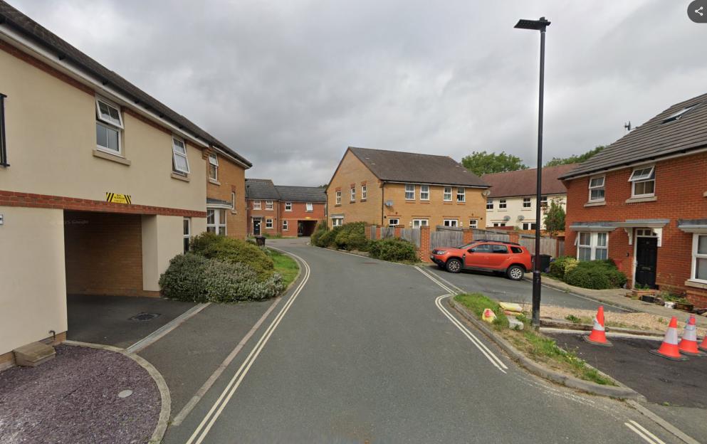 A residential street with two-story brick and beige houses, an orange SUV parked on the side of the road, and an overcast sky.