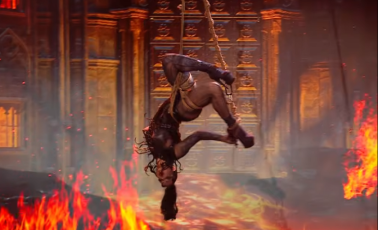 A woman hangs upside down from a rope, surrounded by flames, with a large, ornate building in the background.