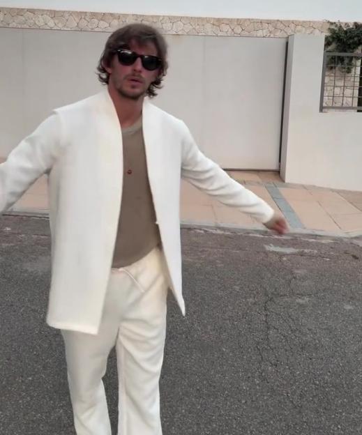 A man in a white blazer, white pants, and sunglasses stands with arms outstretched on a paved surface.