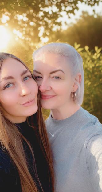 A close-up selfie of a woman with long brown hair and a woman with short blonde hair, smiling at the camera with sunlight behind them.