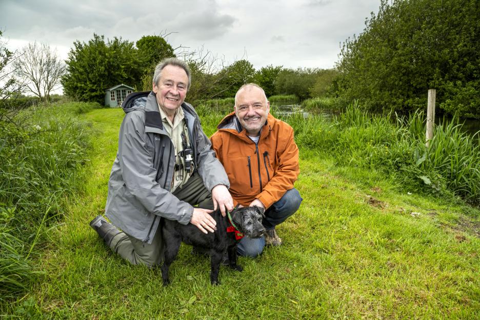 Paul Whitehouse, Bob Mortimer, and Ted the dog kneeling in a grassy field next to a river.