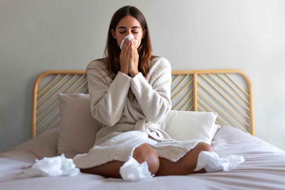 Young woman blowing nose with a tissue while sitting on a bed.