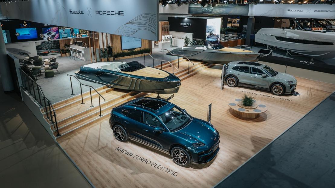 Overhead shot of a Porsche exhibit with two electric SUVs and three yachts, two labeled Invictus Yachts, under a banner for "Frauscher x Porsche."