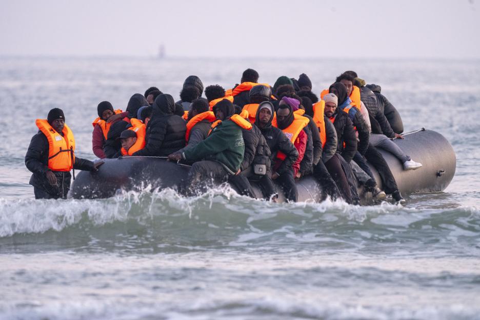 A group of migrants, wearing orange life vests, packed into a small inflatable boat in the water.