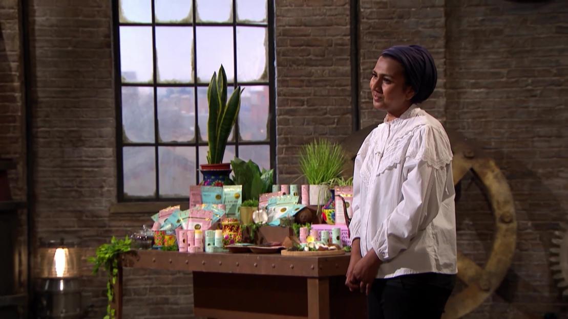 A woman in a white blouse and blue head covering stands next to a table displaying various packaged products.