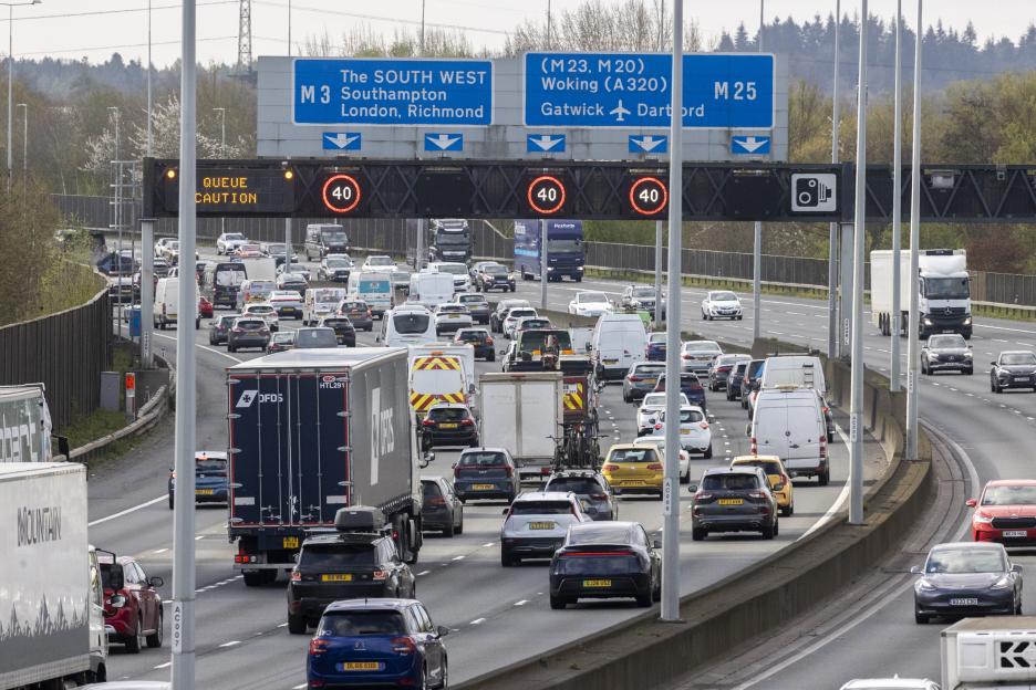 Heavy traffic on the M25 in Egham, UK, with a digital sign showing "QUEUE CAUTION" and a 40 mph speed limit.
