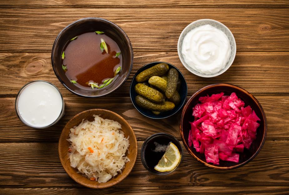 Assorted fermented foods and drinks including kimchi, pickles, sauerkraut, miso soup, kombucha, yogurt, and kefir on a wooden background.