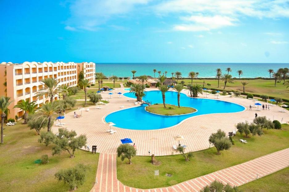 An aerial view of the Nour Palace Resort in Mahdia, Tunisia, showing the hotel building, a large swimming pool, palm trees, and the ocean in the background.