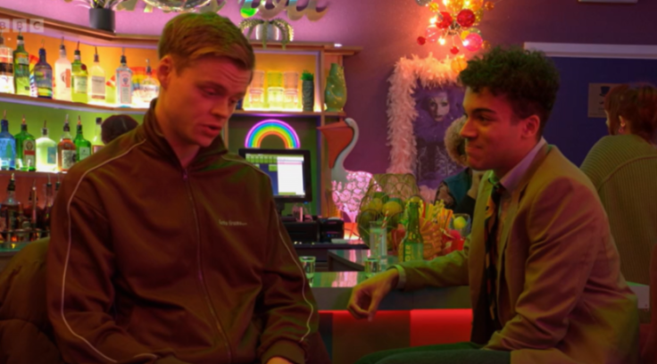 Two men sitting at a bar with colorful lights and a rainbow sign in the background.