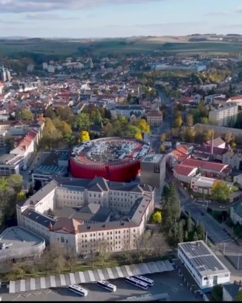 Aerial view of the Horacka Multifunctional Arena in Jihlava, a red stadium with a running track on its roof, surrounded by a city with buildings and green spaces.