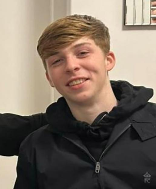 A young man with light brown hair smiles while wearing a black jacket.