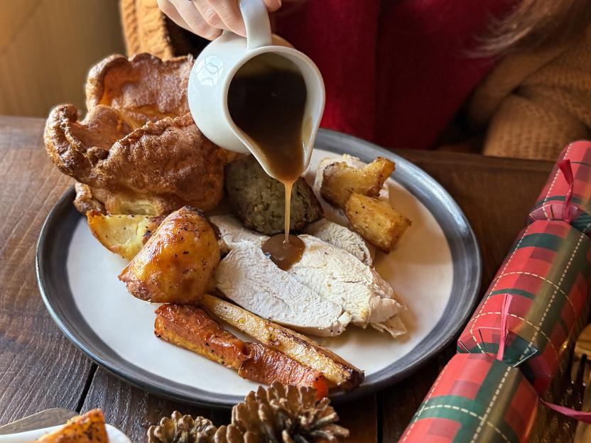 A person pours gravy from a white pitcher onto a plate of Christmas roast dinner, which includes sliced chicken, roasted potatoes and carrots, Yorkshire pudding, and stuffing, next to a red and green plaid Christmas cracker.