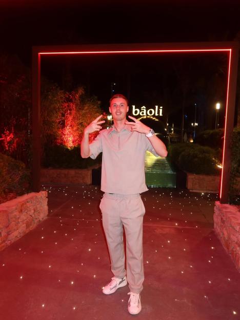 Cole Palmer standing in front of a red neon frame with "bâoli" written above him, giving peace signs.