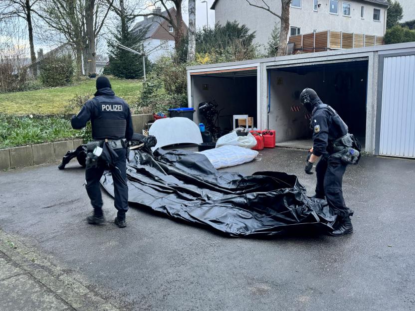 Two police officers in tactical gear examine a large black tarp and other items in front of open garages.