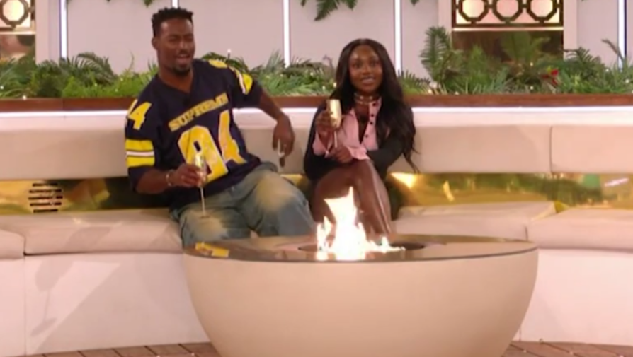 A man and a woman in a Love Island setting, drinking champagne next to a fire pit.
